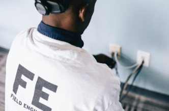 Electrical safety media - field engineer in white FE shirt kneeling testing wall sockets with cables